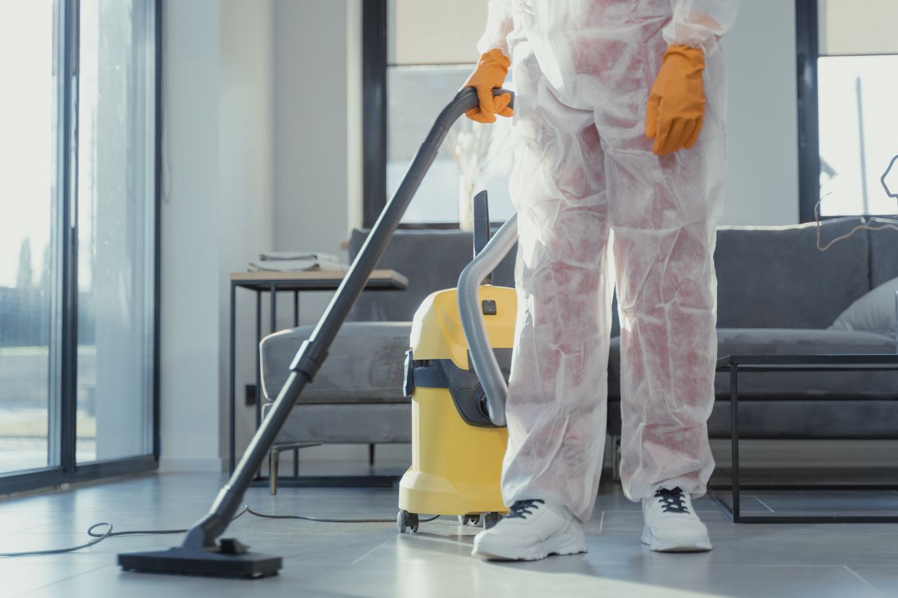 A cleaner wearing protective coveralls and gloves, vacuuming a contemporary living room.