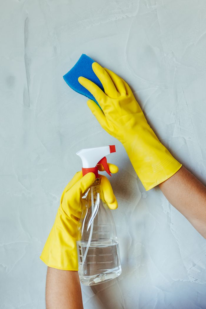 Mains gantées de jaune nettoyant un mur avec une éponge et un flacon pulvérisateur.